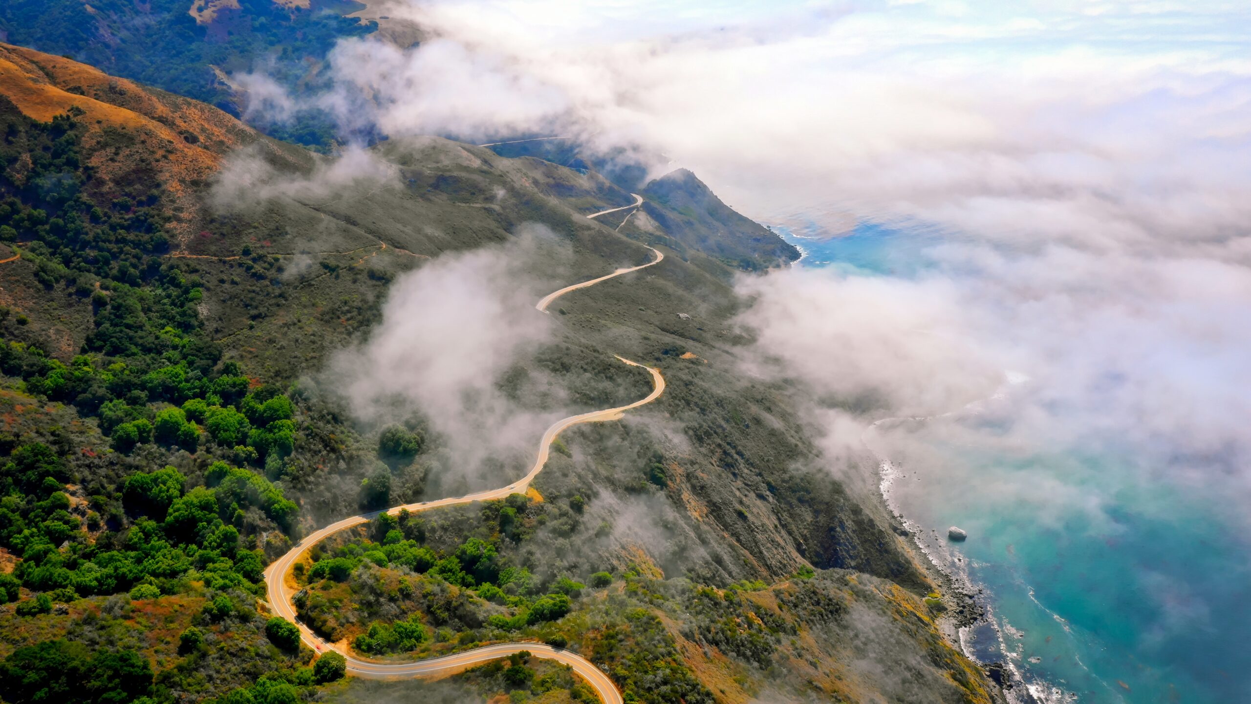 An aerial shot of beautiful green hills and a curvy road going along on the edge and amazing sea with clouds