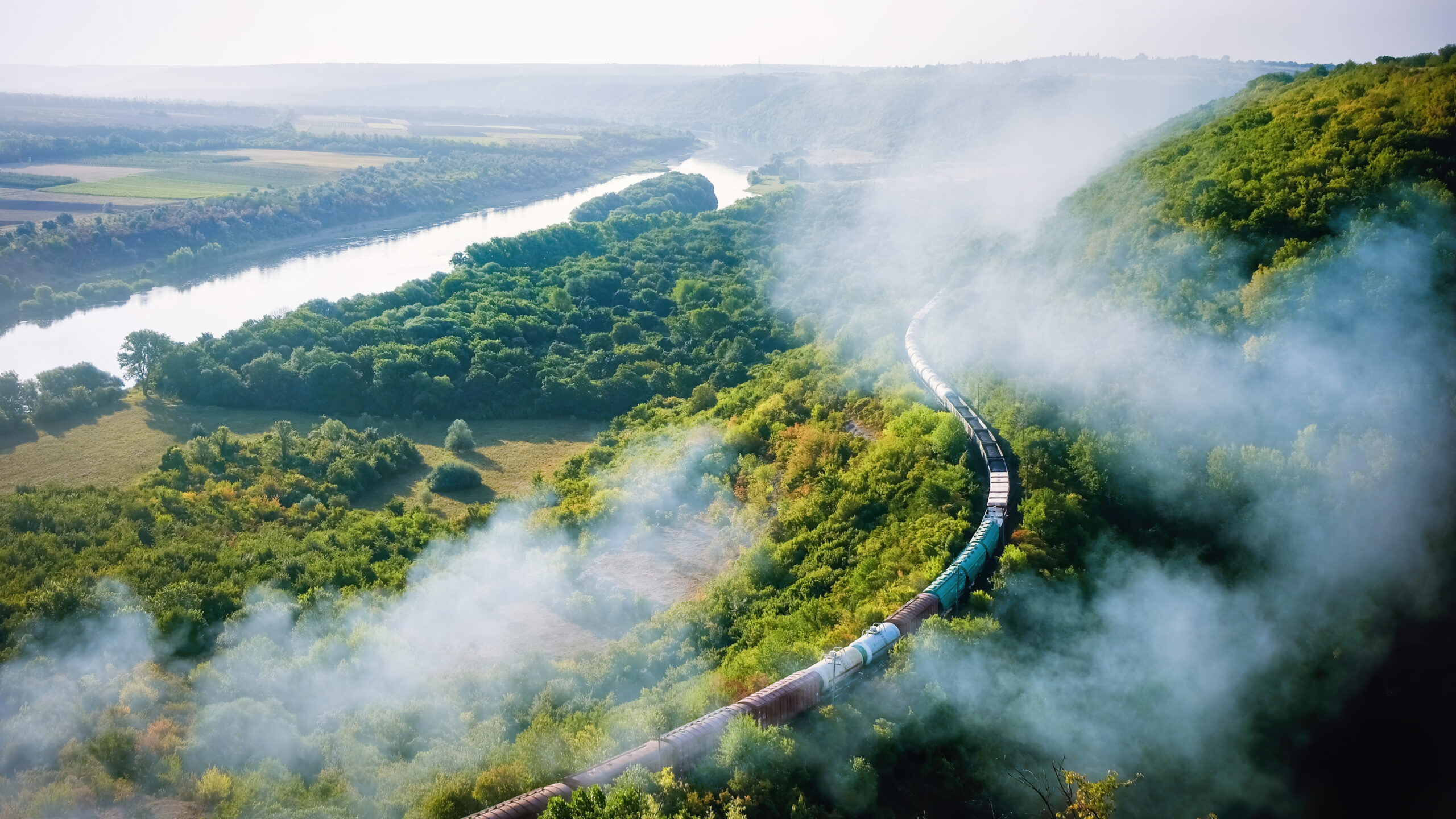 Moving train on railway with high column of smoke, flowing river, hills and railway on the foreground, fields on the other shore in Moldova